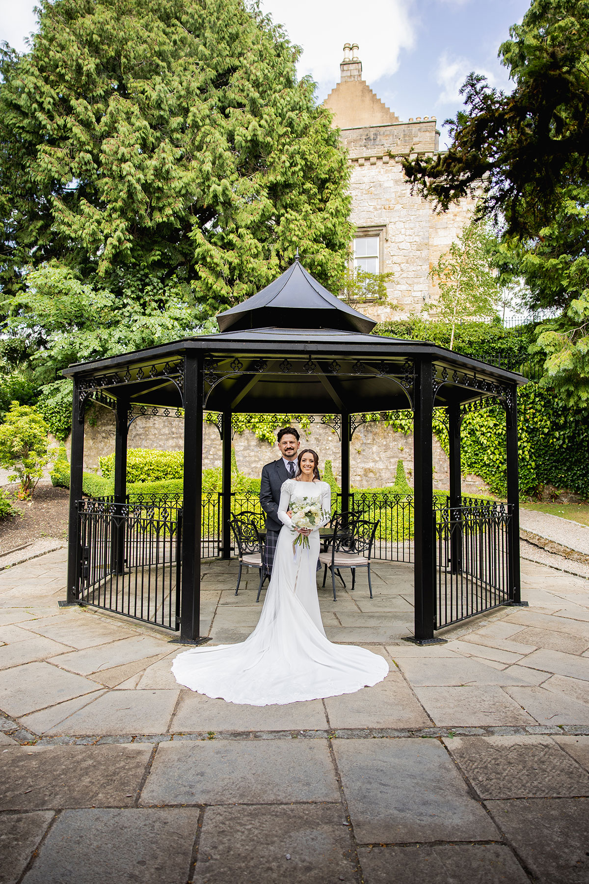 bride and groom stand smiling under a black wrought-iron gazebo in the castle gardens, the bride holding a bouquet of white flowers
