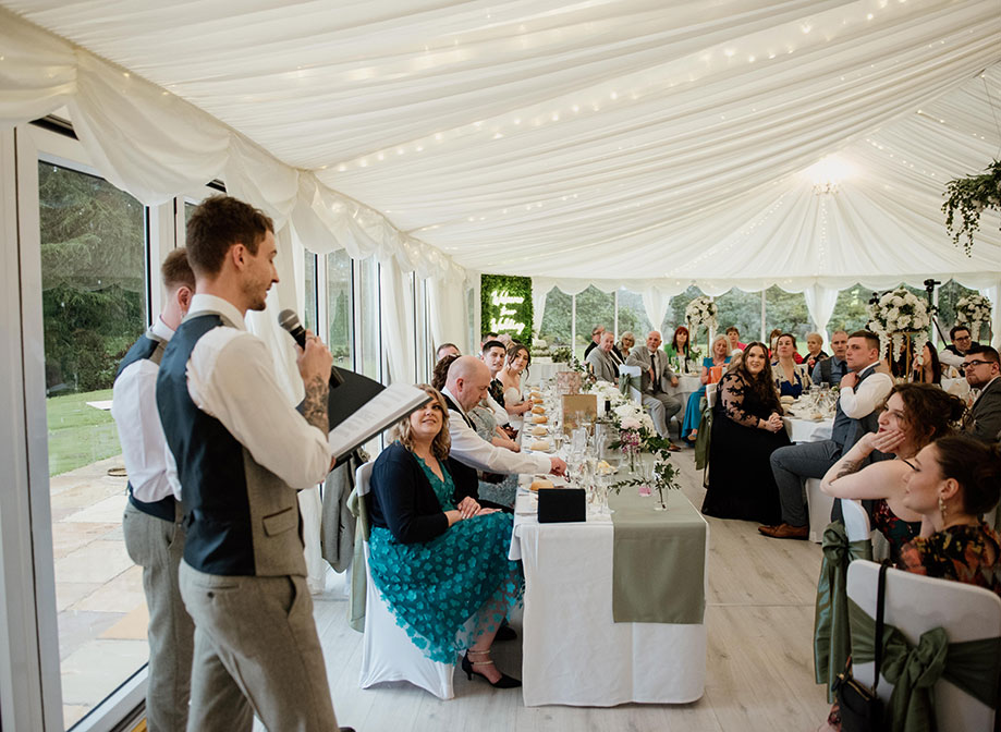 Two men in waistcoats and smart trousers holding microphones give a speech to a crowd