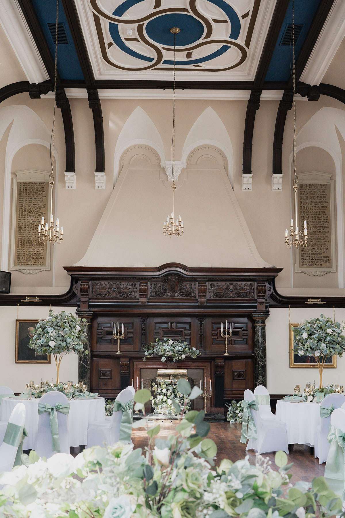 Wide view of the Grand Hall at 1599 at the Royal College set for a wedding reception with mint green chair sashes, floral centrepieces, high ceilings and an ornate carved fireplace
