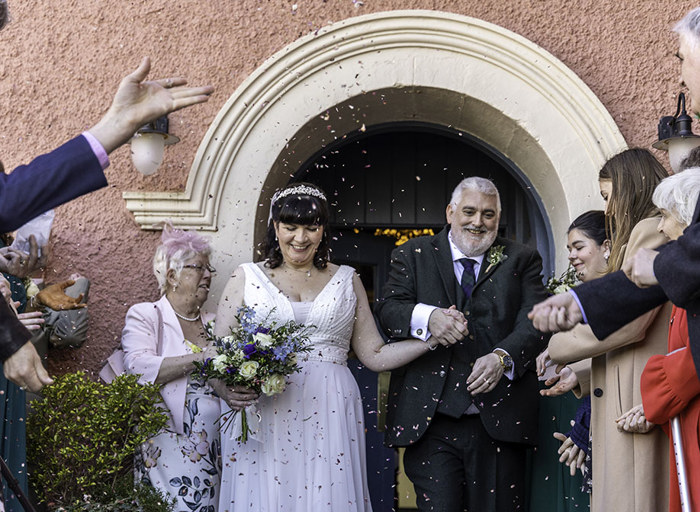 a bride and groom waking through two rows of wedding guests that are throwing confetti. They are outside a pink coloured building.