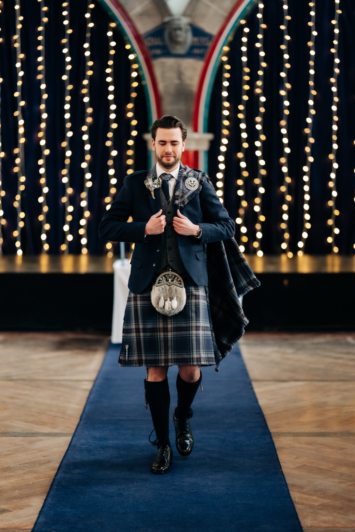 Groom James poses in his tartan kilt and navy jacket beneath fairy lights in Òran Mór's Auditorium before the wedding ceremony.