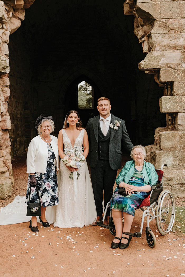 a bride and groom posing for a photo with an elderly lady in a wheelchair and another elderly lady in front of a ruined stone building at Dunglass Estate