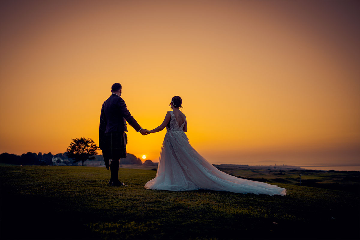 A bride in a dress with a long train and a groom wearing a kilt stand holding hands looking at a sunset which fills most of the image with yellow light