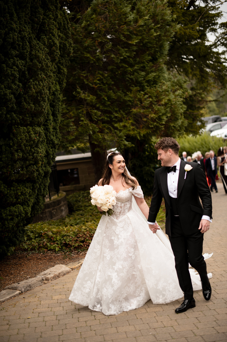 Bride carrying a white rose bouquet and groom in a black tuxedo walking together through the grounds of Crossbasket Castle.