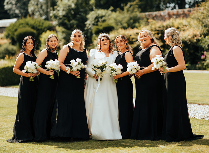 Six bridesmaids wearing black dresses stand on either side of a bride wearing a white wedding dress and hairband