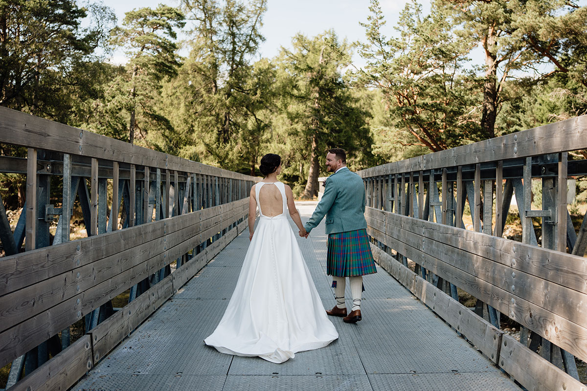 bride and groom holding hands while walking across a wooden bridge in the Cairngorms after their wedding ceremony