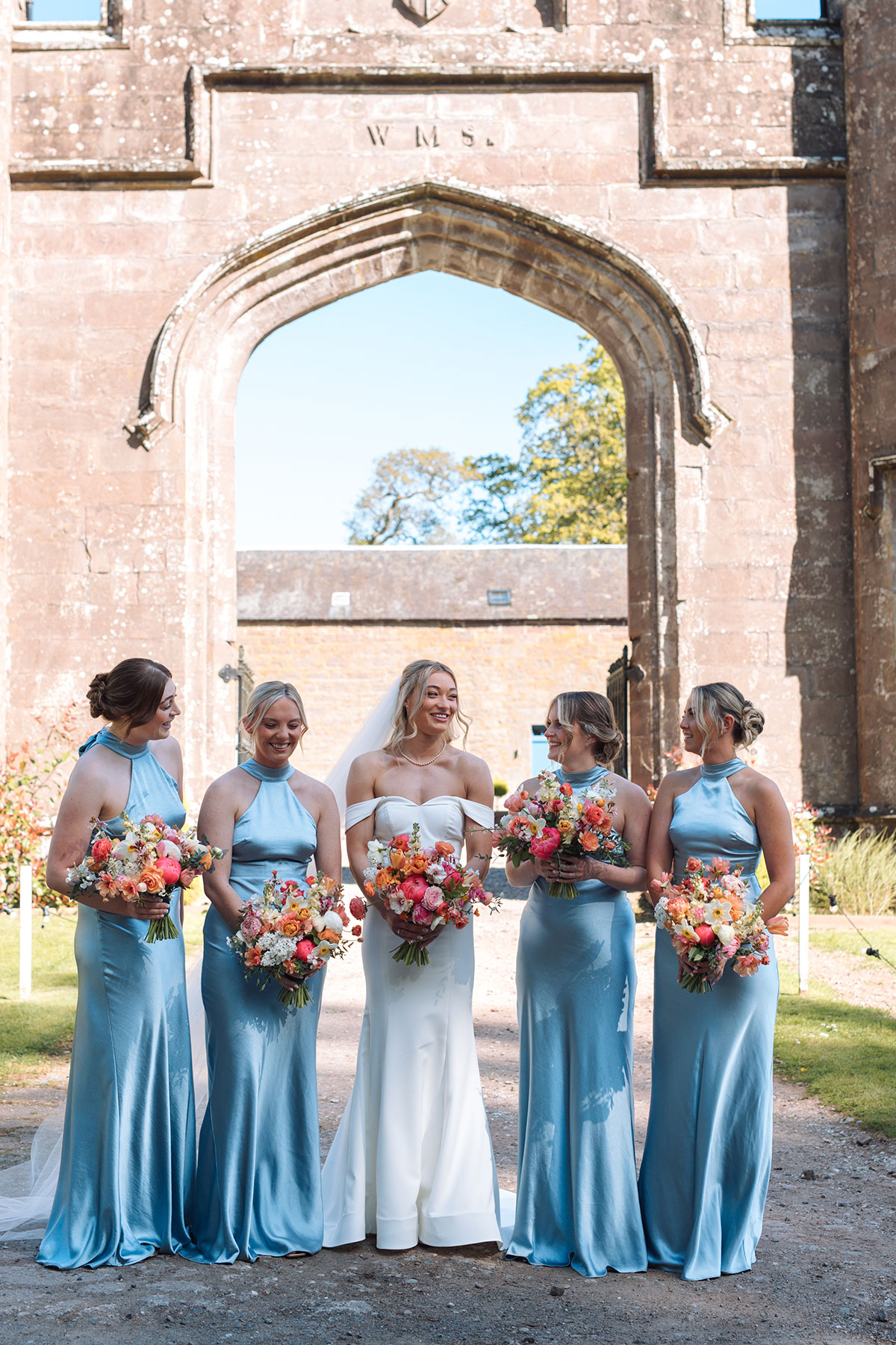 Bride and bridesmaids in blue dresses holding colourful bouquets outside Rosebery Estate archway