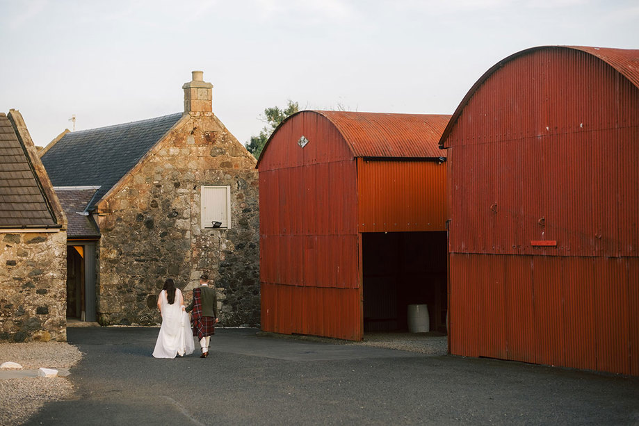 A couple walk away from the camera in the distance past red barns and stone buildings 