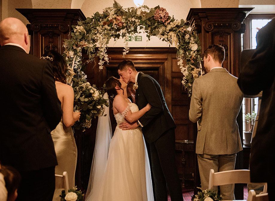 A bride and groom kiss underneath a flower arch
