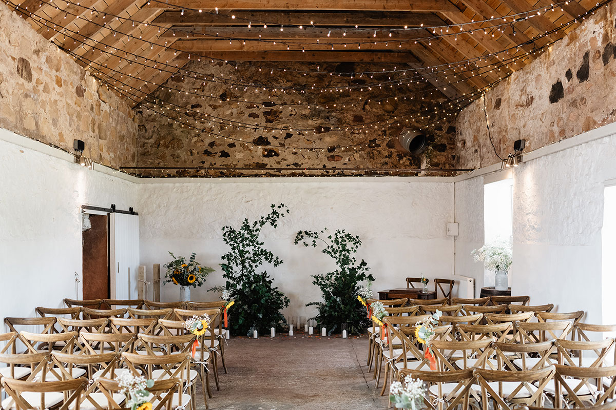 Rustic ceremony room with exposed stone walls, wooden beams and strings of fairy lights, set with wooden chairs and greenery at the aisle end