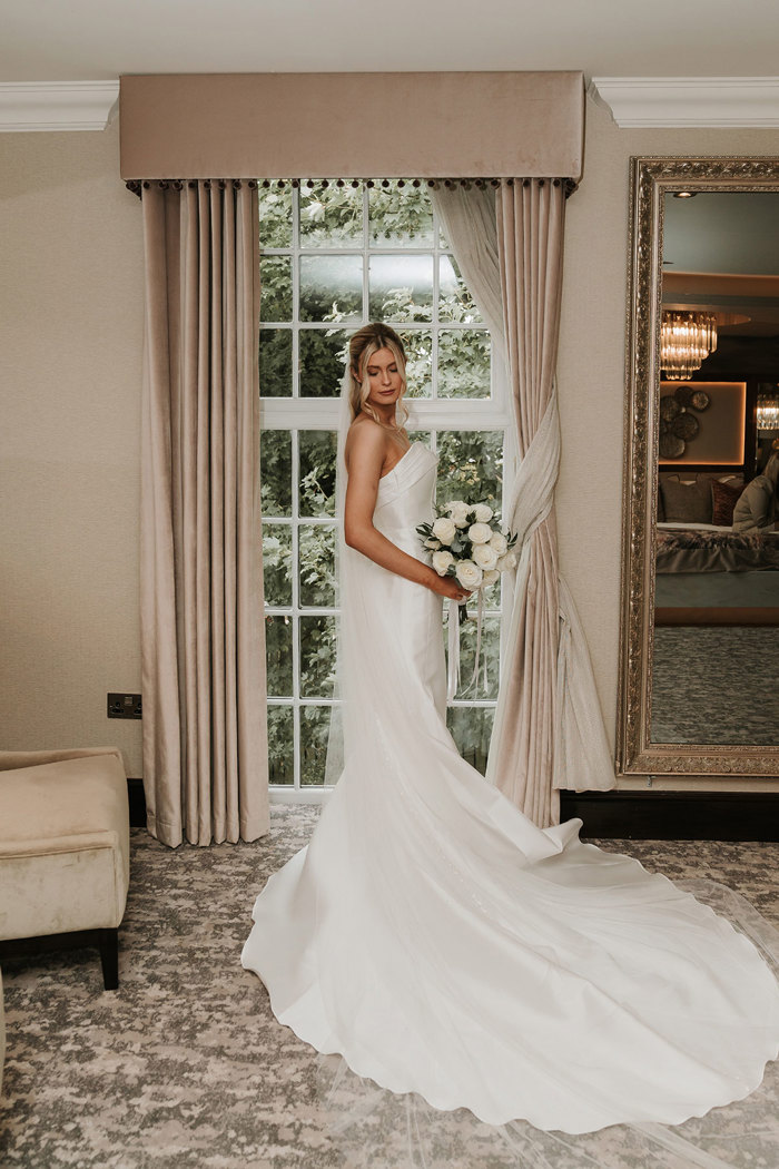 a bride holding a bouquet of white roses posing by a window in Busby Hotel