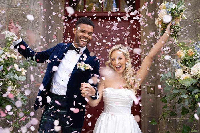 A groom wearing green tartan trousers and a navy velvet suit jacket holds hands with a bride wearing a strapless wedding dress as pink confetti is thrown over them