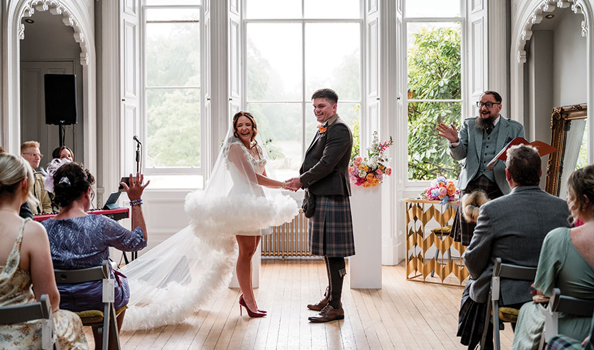 Bride and groom laughing during their ceremony in a bright room with tall windows, the bride wearing a voluminous high-low gown.