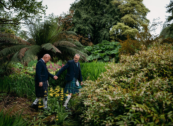 two grooms wearing kilts walking in a verdant green garden setting