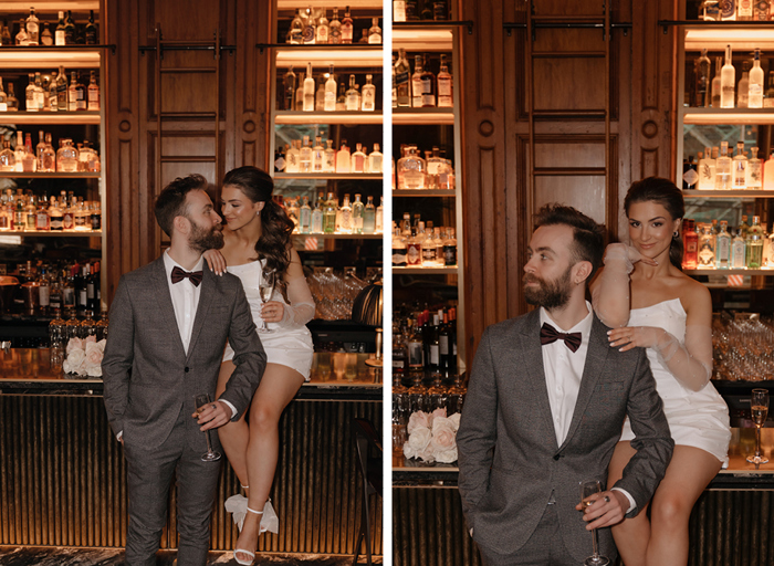A groom stands next to a bar while the bride sits on the bar, both holding a glass of champagne