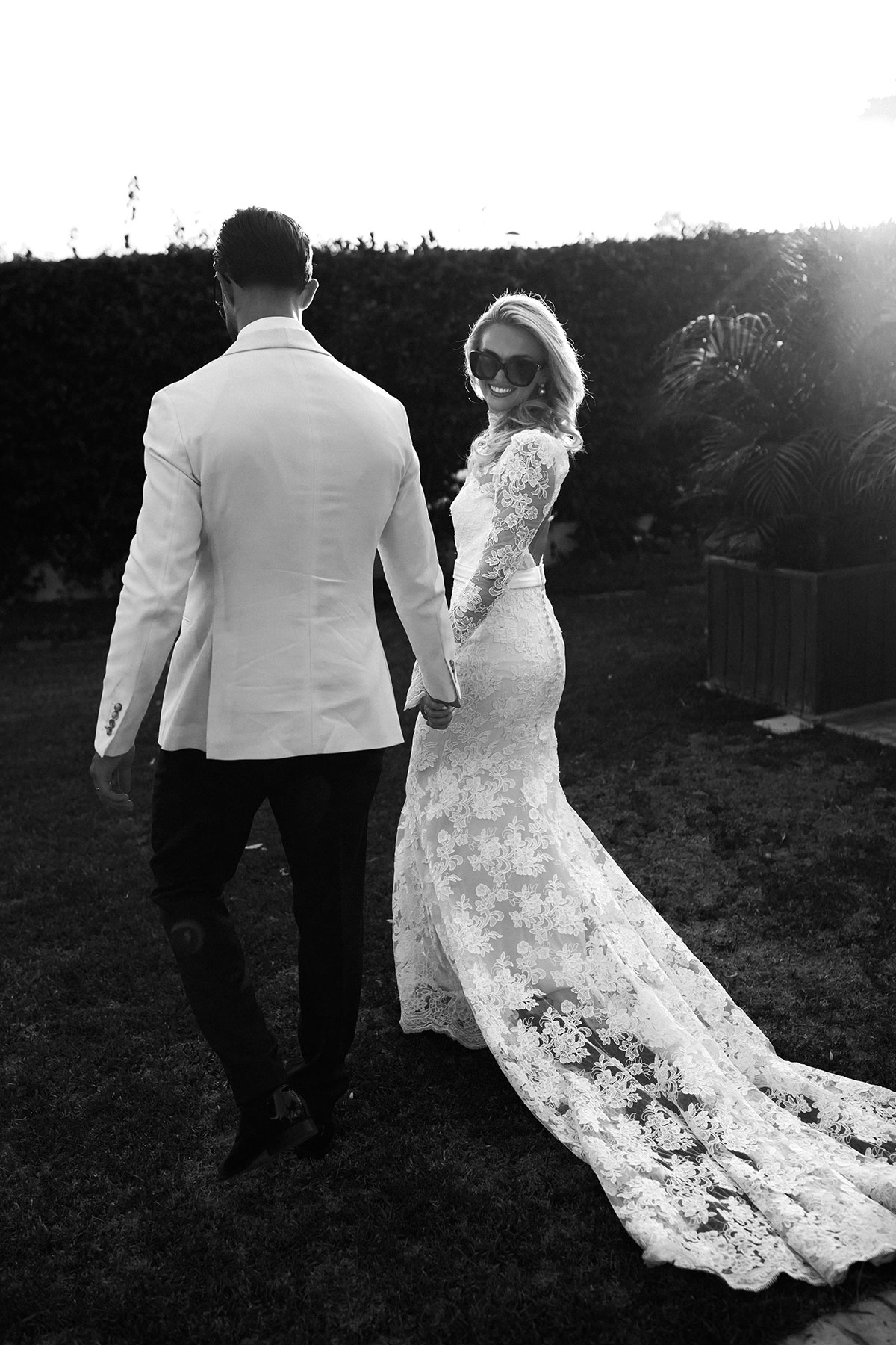 Bride and groom holding hands, bride smiling back over shoulder in lace gown and sunglasses
