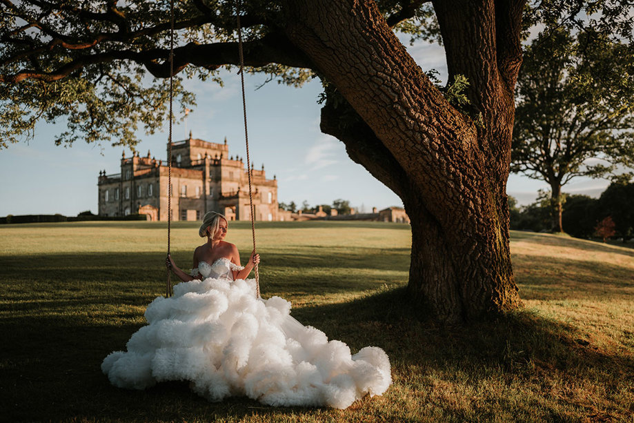 A bride wearing a wedding dress with a voluminous tulle skirt sitting on a swing attached to a tree with a country house in the background