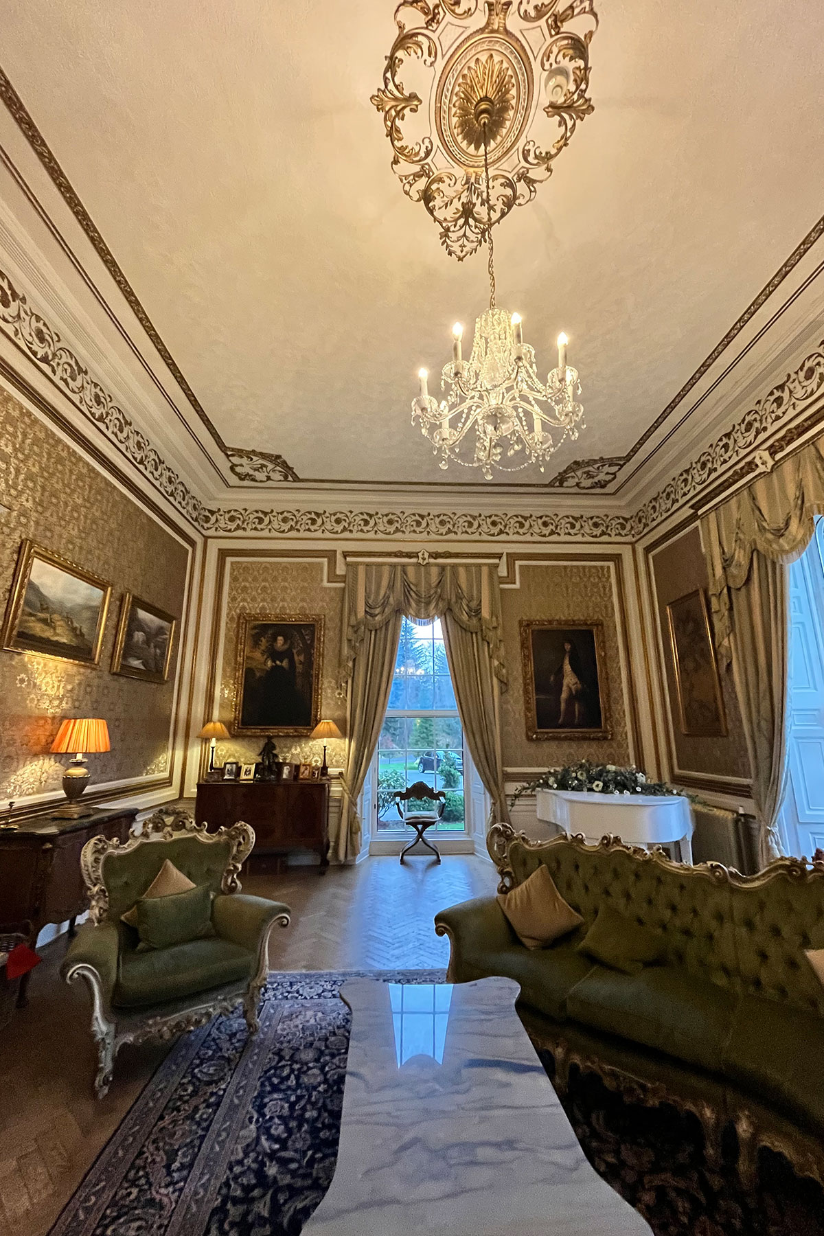 Luxurious castle drawing room with chandeliers, ornate gold decor, green velvet sofas and framed portraits on the walls
