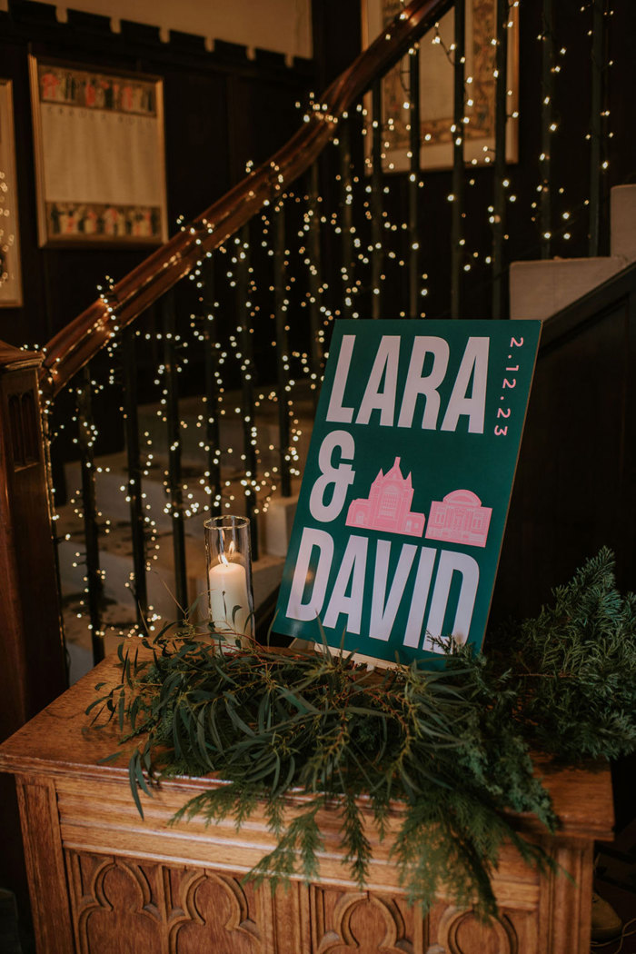 A green sign that reads 'Lara & David 2.12.23' sitting on a wooden table covered in green foliage and a candle in a glass jar, sitting in front of a staircase wrapped in fairylights