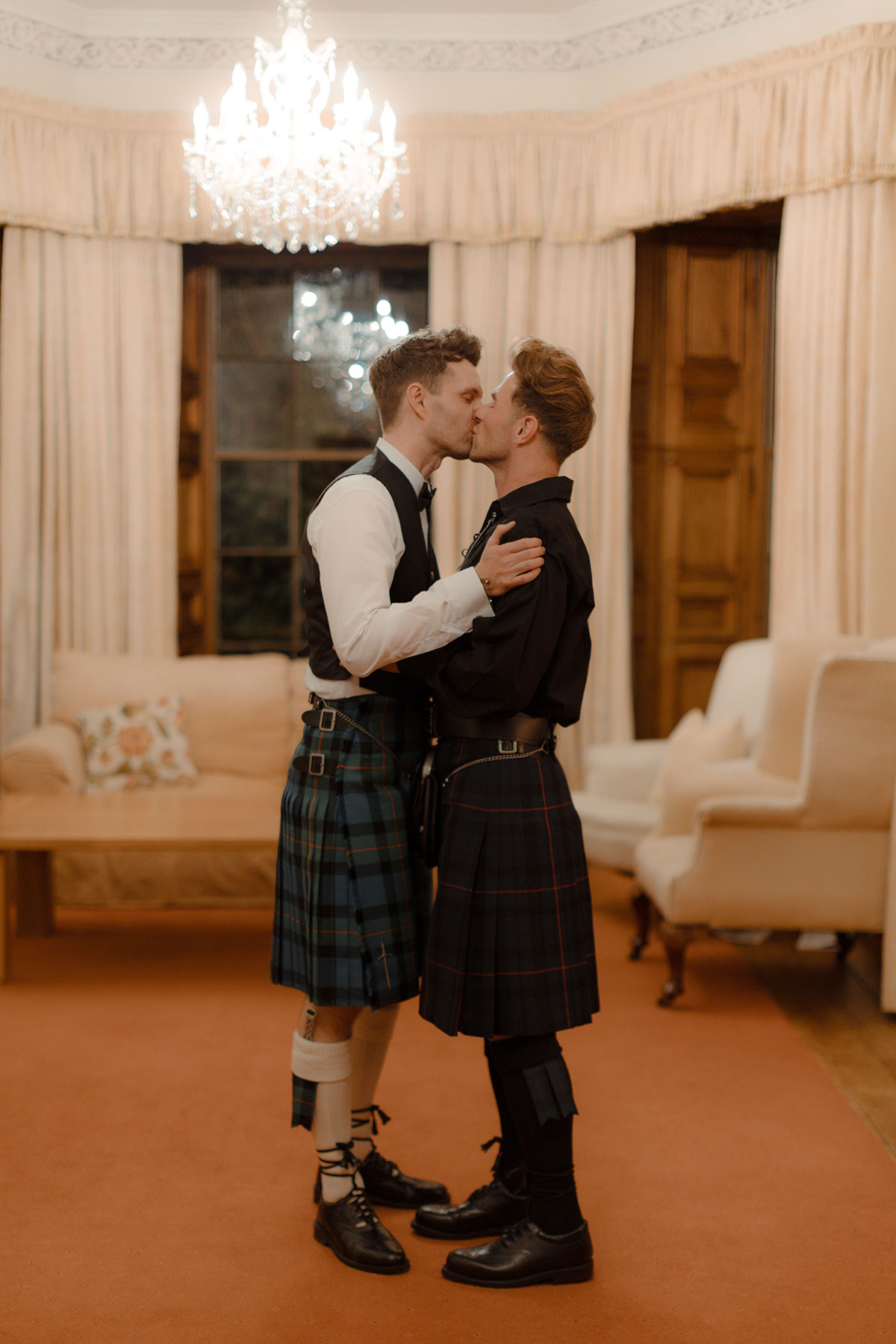 Two grooms share a kiss indoors at Newhall Estate, both wearing Gunn tartan kilts during their wedding reception