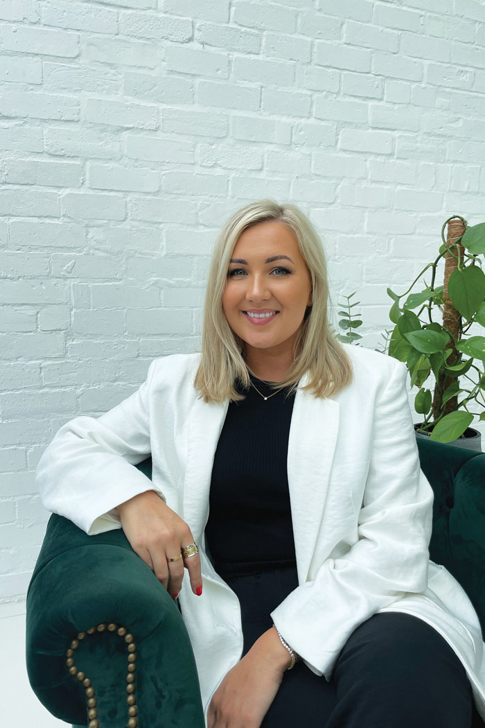 tan woman with shoulder-length blonde hair wears a white blazer and sits in front of white brick wall