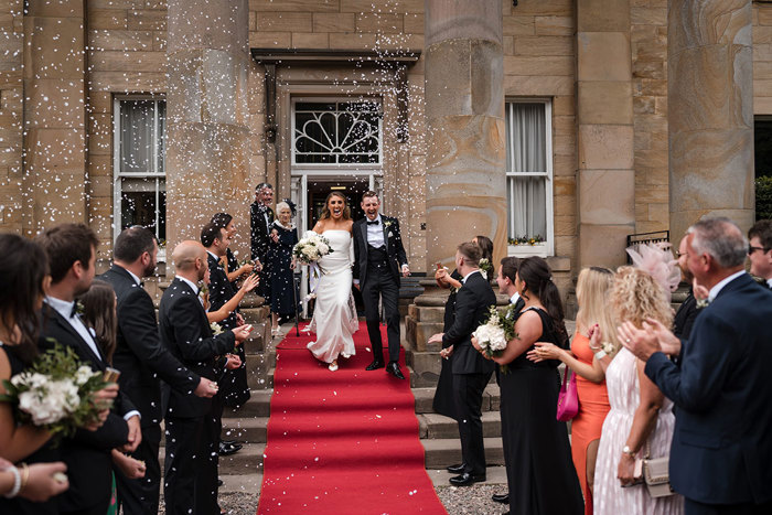 Bride and groom walk down red carpet following their ceremony to guests cheering and throwing confetti