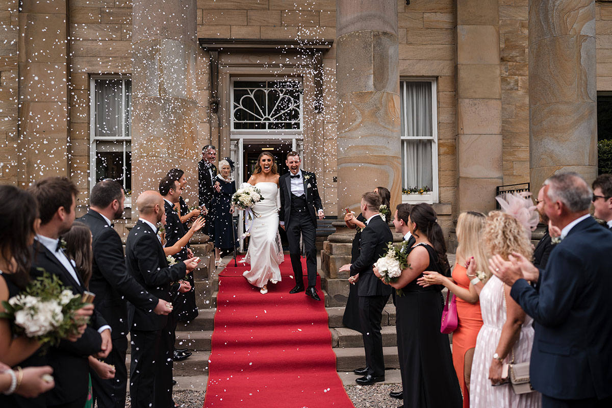 Bride and groom walk down red carpet following their ceremony to guests cheering and throwing confetti