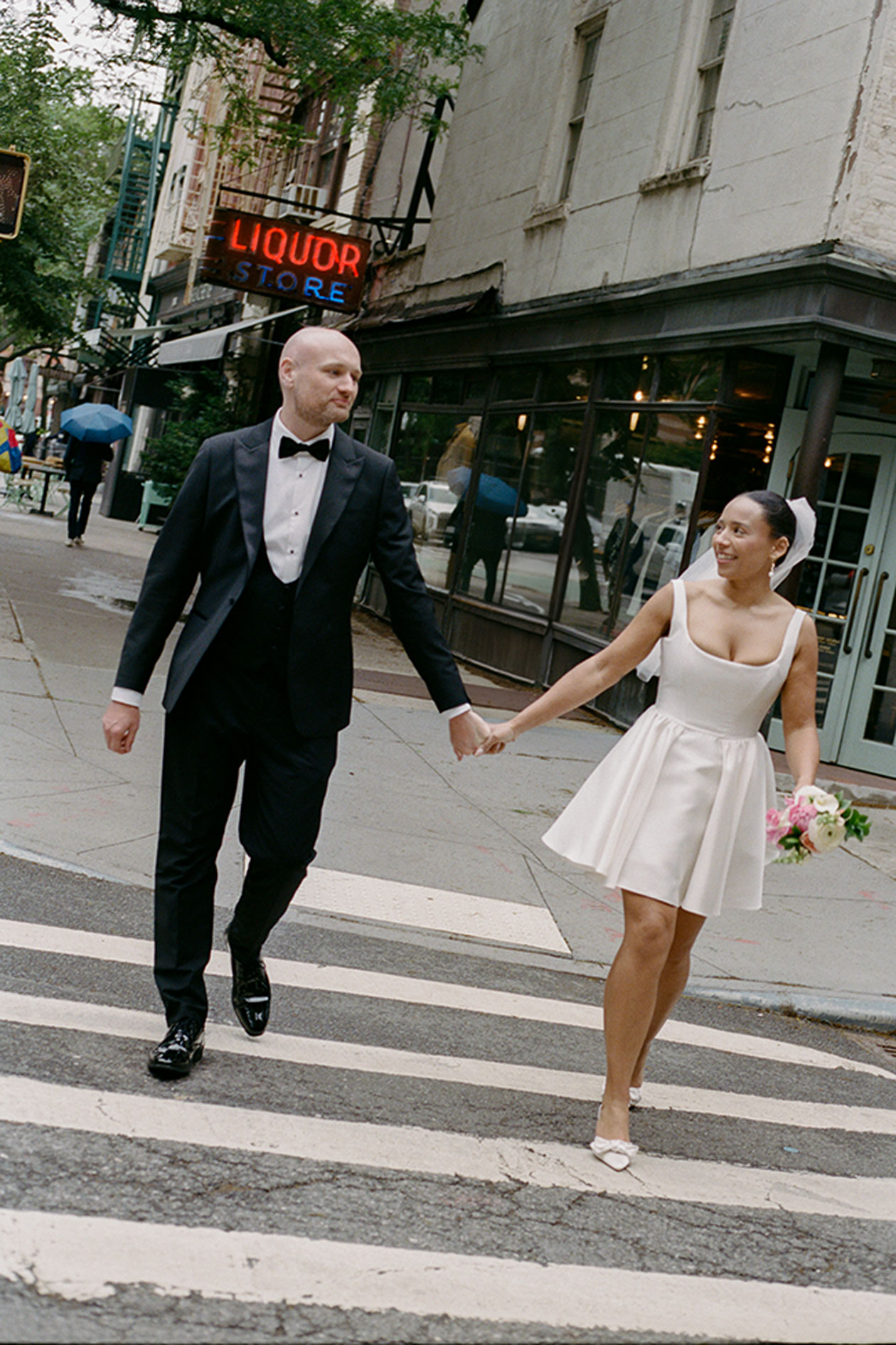 Bride and groom crossing New York street holding hands after City Hall elopement liquor store sign in background