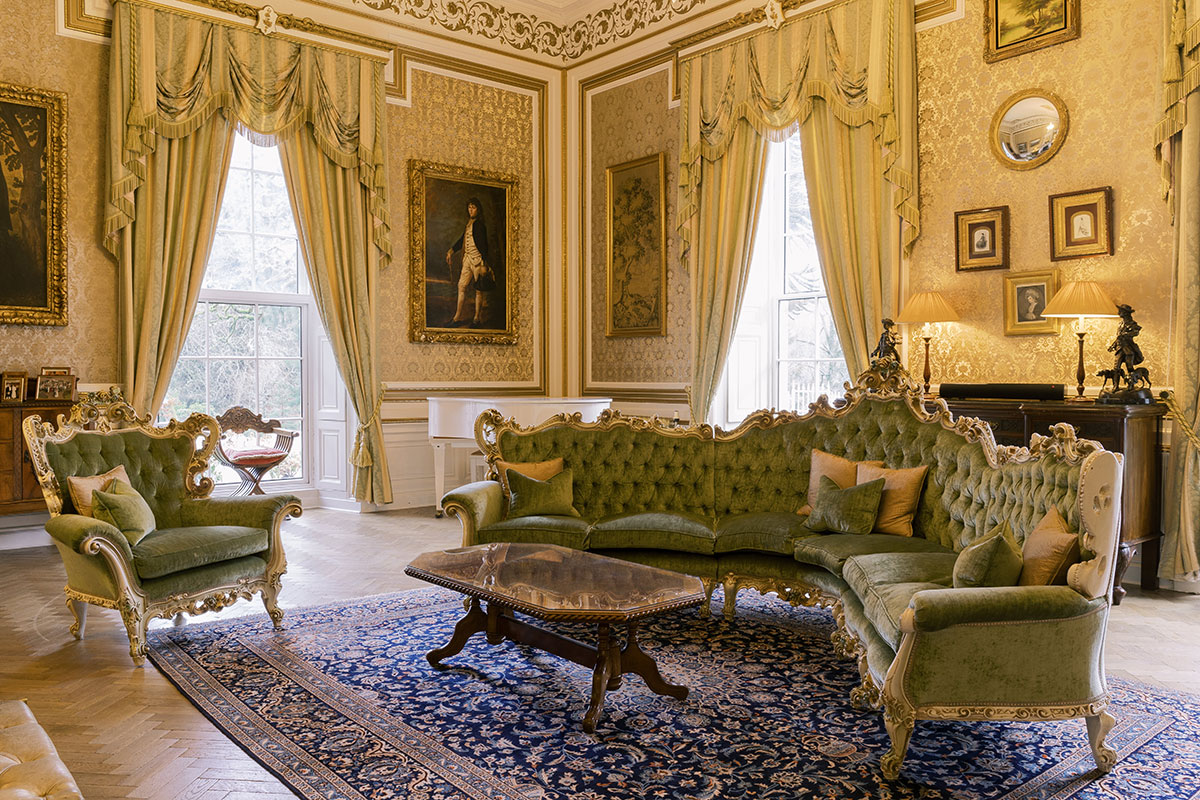 Luxurious drawing room with gold wallpaper, ornate green velvet furniture, framed portraits, and tall windows with draped curtains