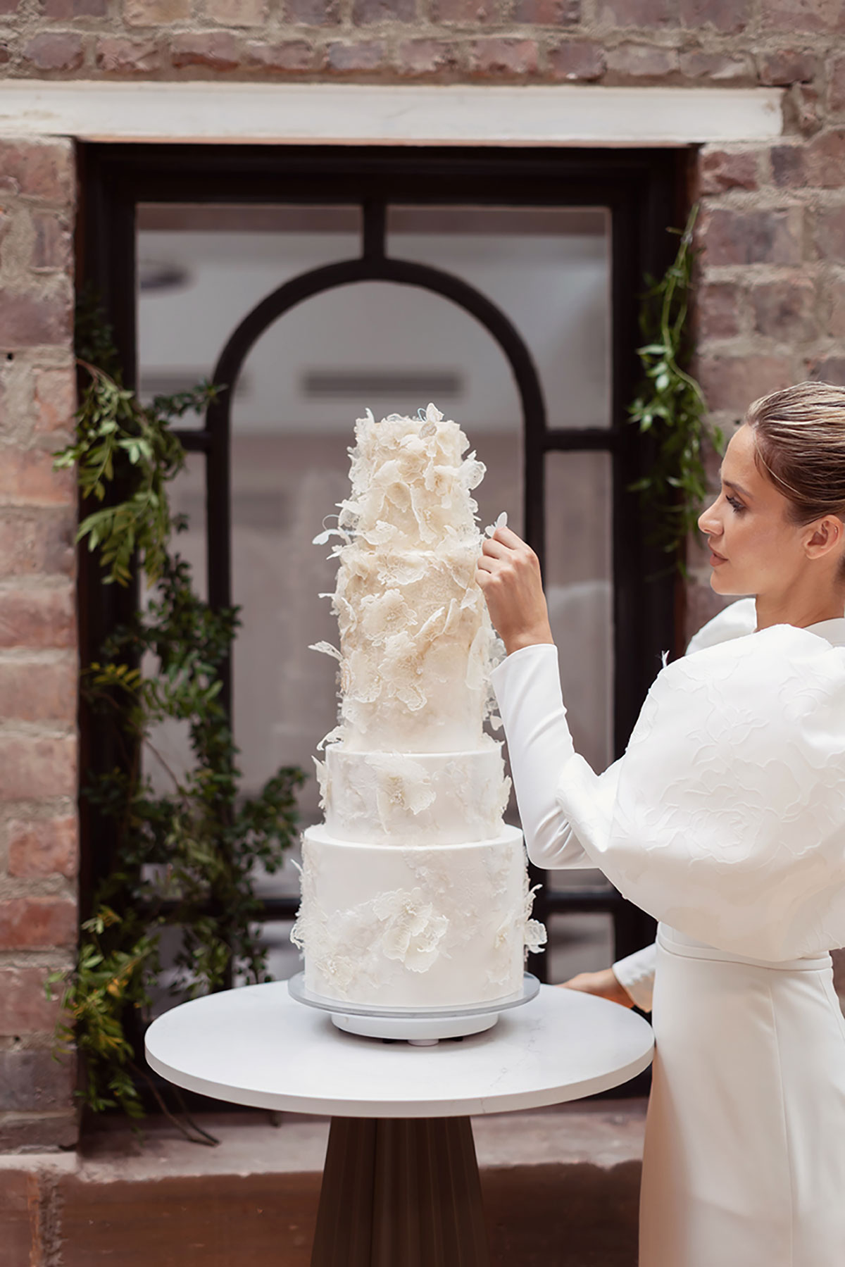 Bride in puff-sleeve gown adjusting tall tiered wedding cake with floral detailing