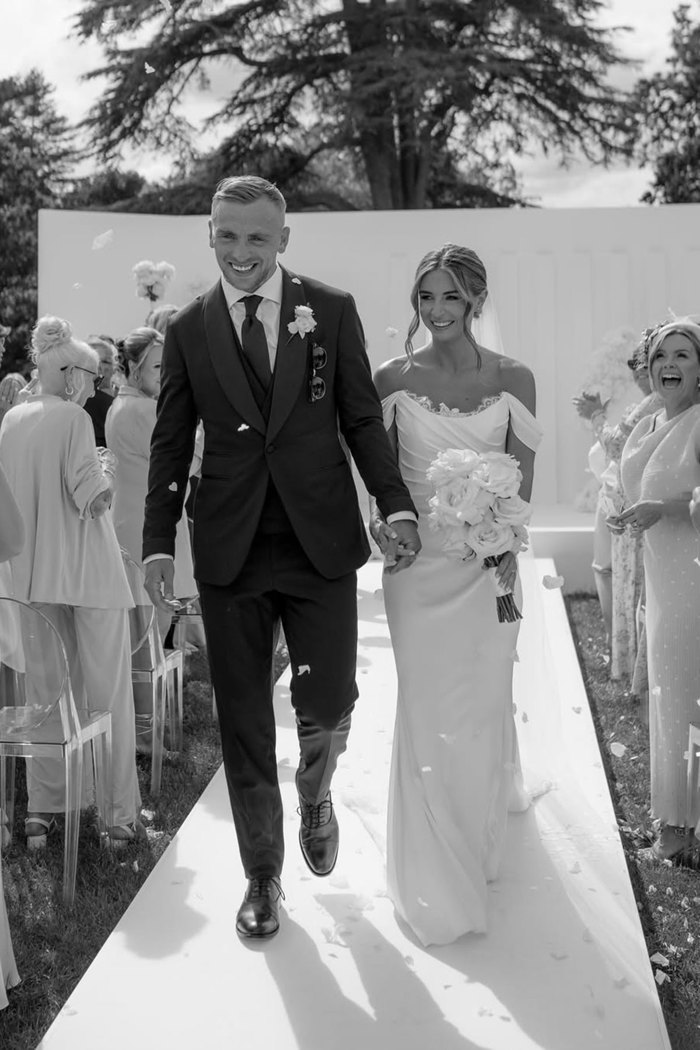 black and white image of a couple walking up the aisle, holding hands as flower petals fall around them, wearing a tux and an off the shoulder wedding dress
