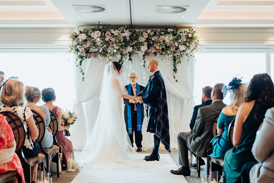 a bride and groom holding hands standing at the top of the aisle at Seamill Hydro during a wedding ceremony. They are standing below a pink and white hanging floral installation as a minister conducts the ceremony and guests watch on
