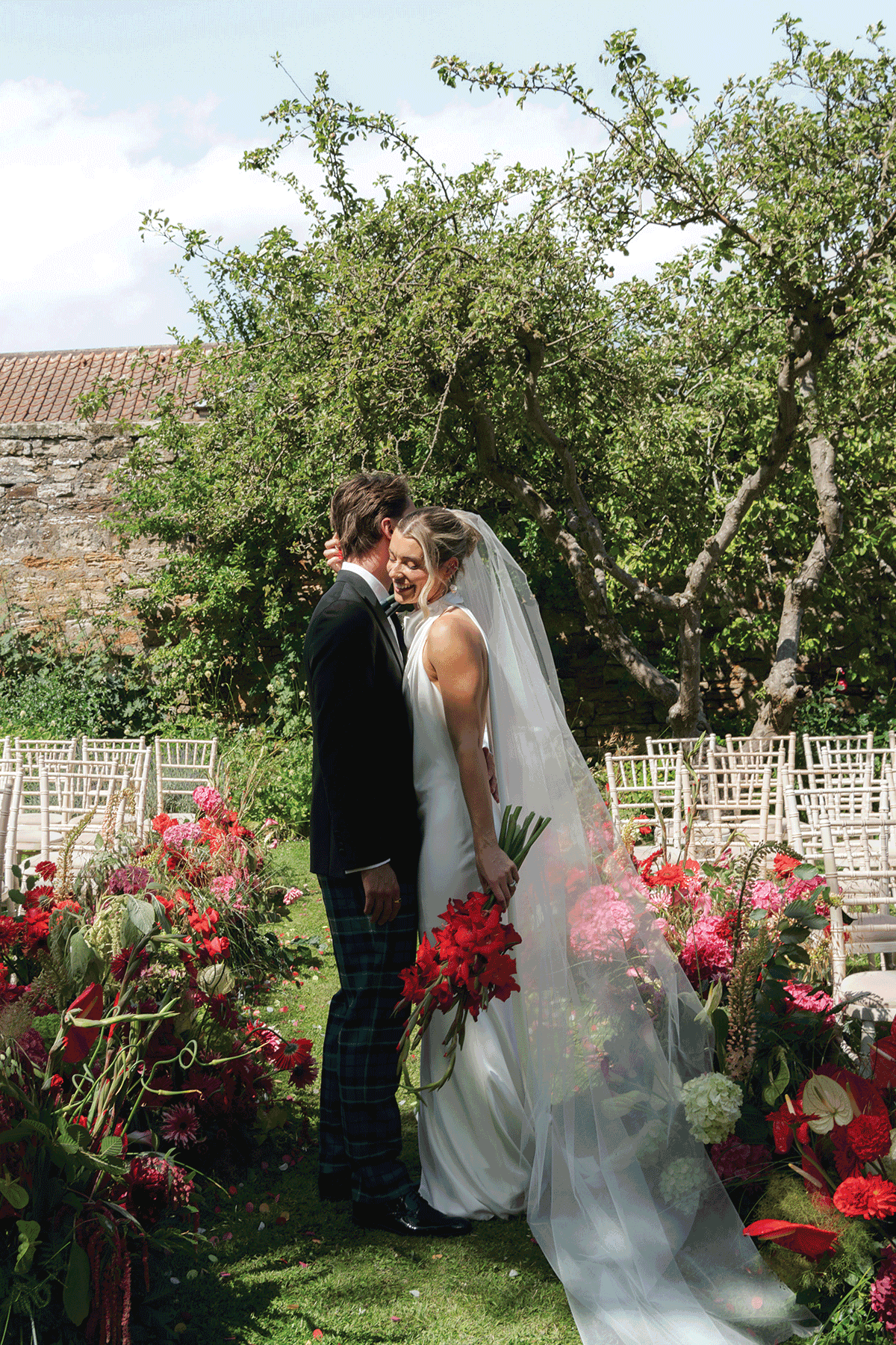Bride and groom embracing in a colourful outdoor wedding ceremony setup filled with bold red and pink florals in a Scottish garden venue