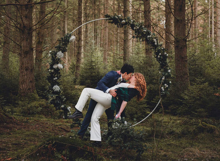 Two brides do a dipped kiss in front of a floral moon gate in a forest