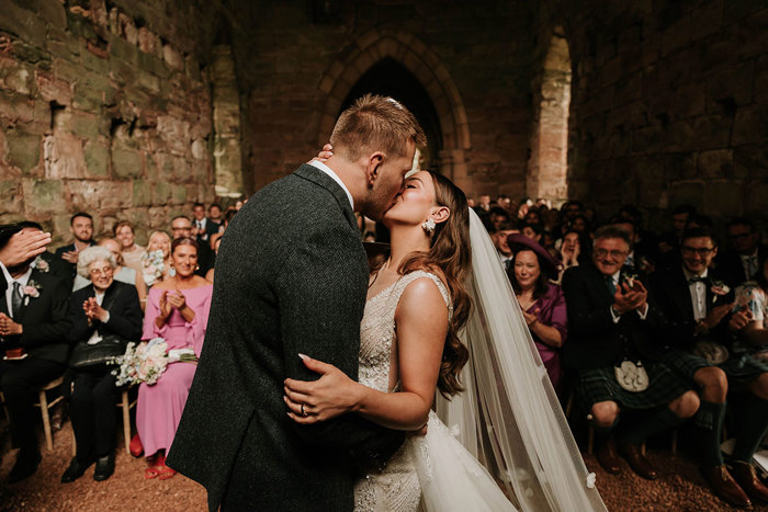 a bride and groom Matt Fagerson kissing after their wedding ceremony in Dunglass Collegiate Church as rows of seated guests clap