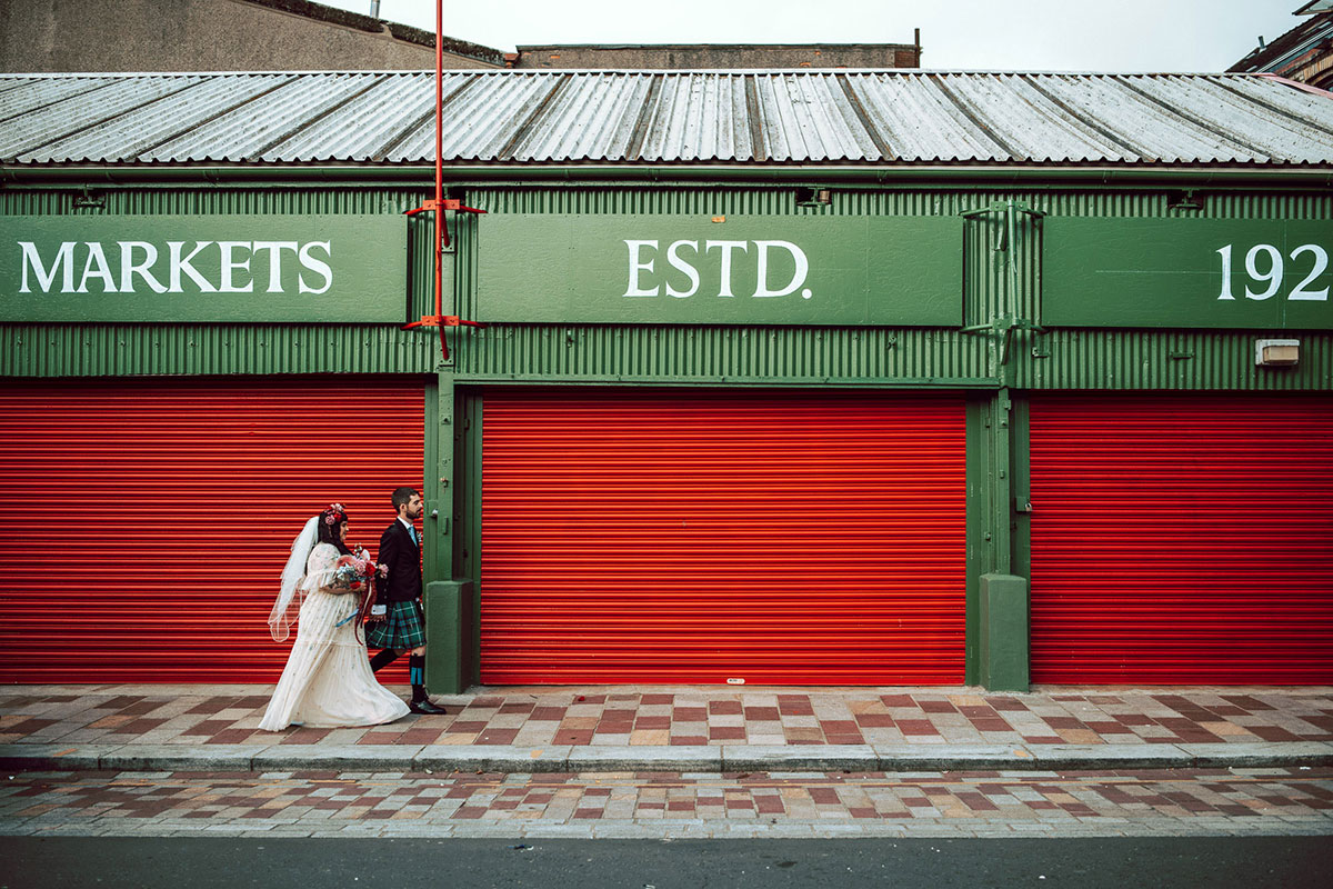 bride and groom walk hand in hand at the barras in glasgow in front of a green sign that reads 'markets'