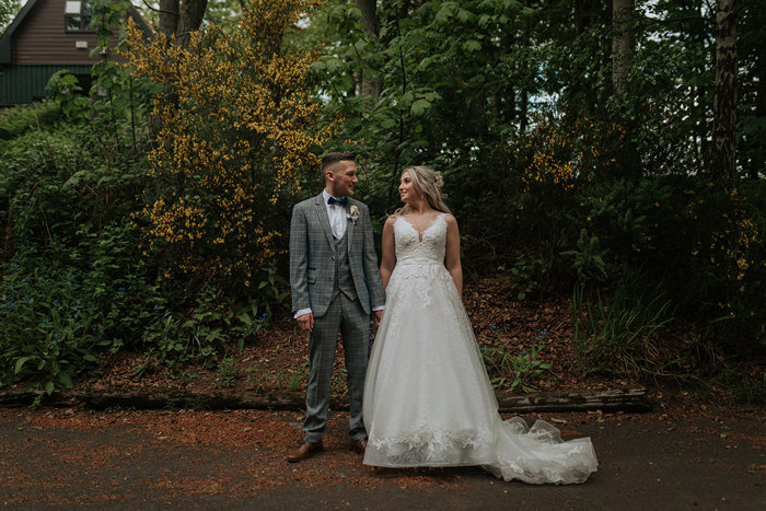 A Bride And Groom Look At Each Other While Standing Against A Mixture Of Trees And Green Hedges