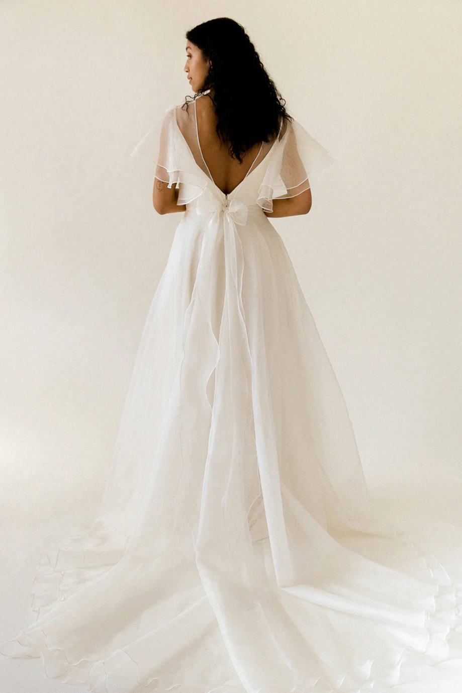 curly dark-haired model wears floaty white wedding dress with sheer layer over bodice providing fluttery sleeves and a small bow at the back of waist trailing down along with the skirt