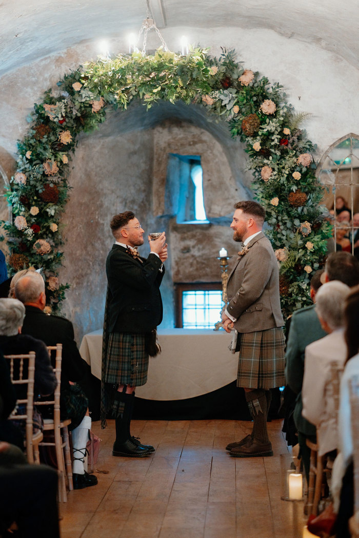 two grooms wearing kilts stand and face each other below a large flower and foliage arch during a wedding ceremony at Neidpath Castle. One of them is drinking from a silver quaich.