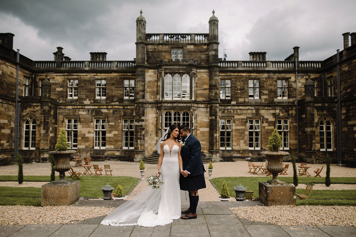 bride wearing a lace wedding gown and holding a bouquet holds husband's hand who is wearing a kilt outside Mar Hall