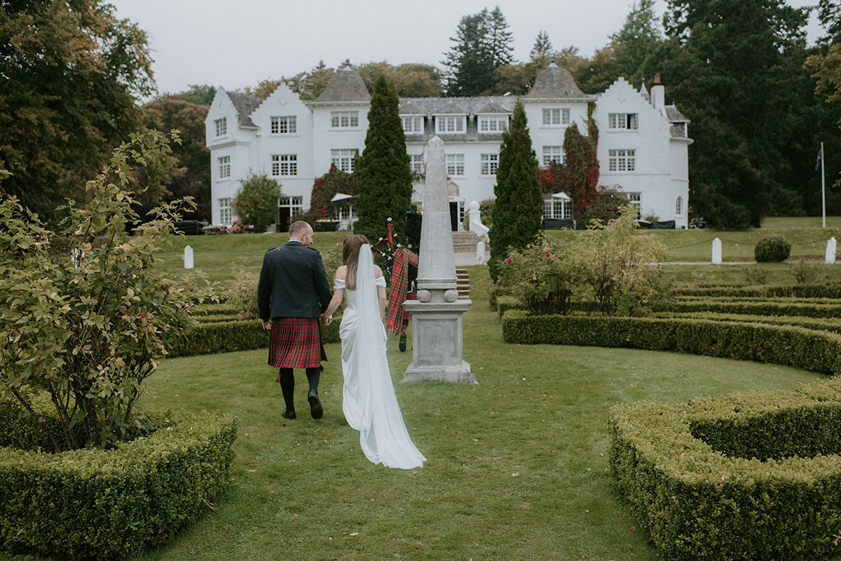 a bride and groom walking behind bagpiper in garden of Achnagairn Castle