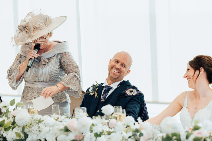 a lady wearing a silver ornate dress and large hat holds a microphone and stands as a bride and groom sit next to her and laugh. There is a row of pink and white flowers in foreground
