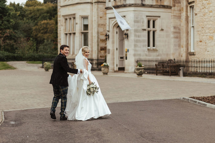 bride and groom smile at camera as he holds the train of her dress walking towards crossbasket castle