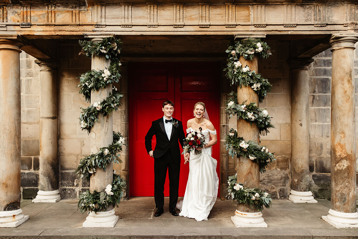 Bride and groom standing beneath festive garlands wrapped around stone columns at Canongate Kirk, in front of red doors.