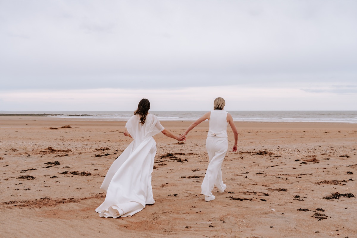 Hannah and Donna walking hand in hand across the beach during their Harvest Moon wedding.