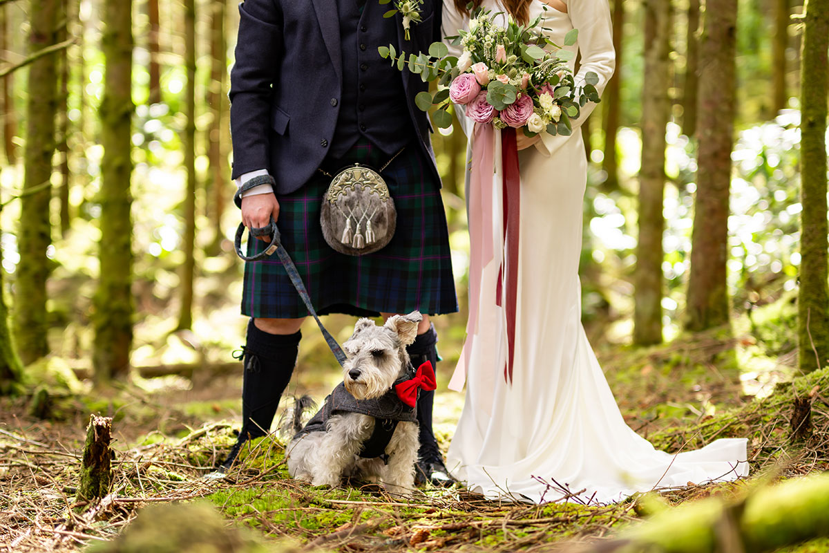 Bride and groom with their schnauzer dog in a Scottish woodland after their Glencoe elopement, with the groom wearing a kilt and the bride holding a pink and white bouquet