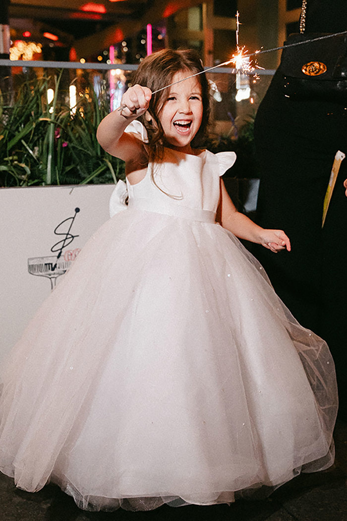 little brunette girl in a poofy white gown with white ribbon tied around the back of waist holds a sparkler in the air and smiles big