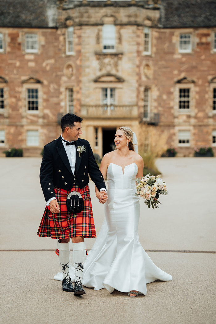 A bride and groom in a kilt holding hands and walking in front of Archerfield House.