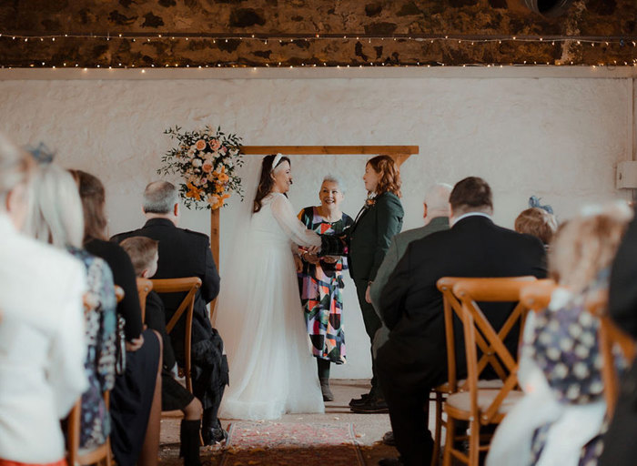 two brides and a wedding celebrant during a wedding ceremony at Cow Shed Crail. They stand below a wooden arch with a flower posy on the top left corner and rows of wedding guests sitting on wooden cross back chairs look on