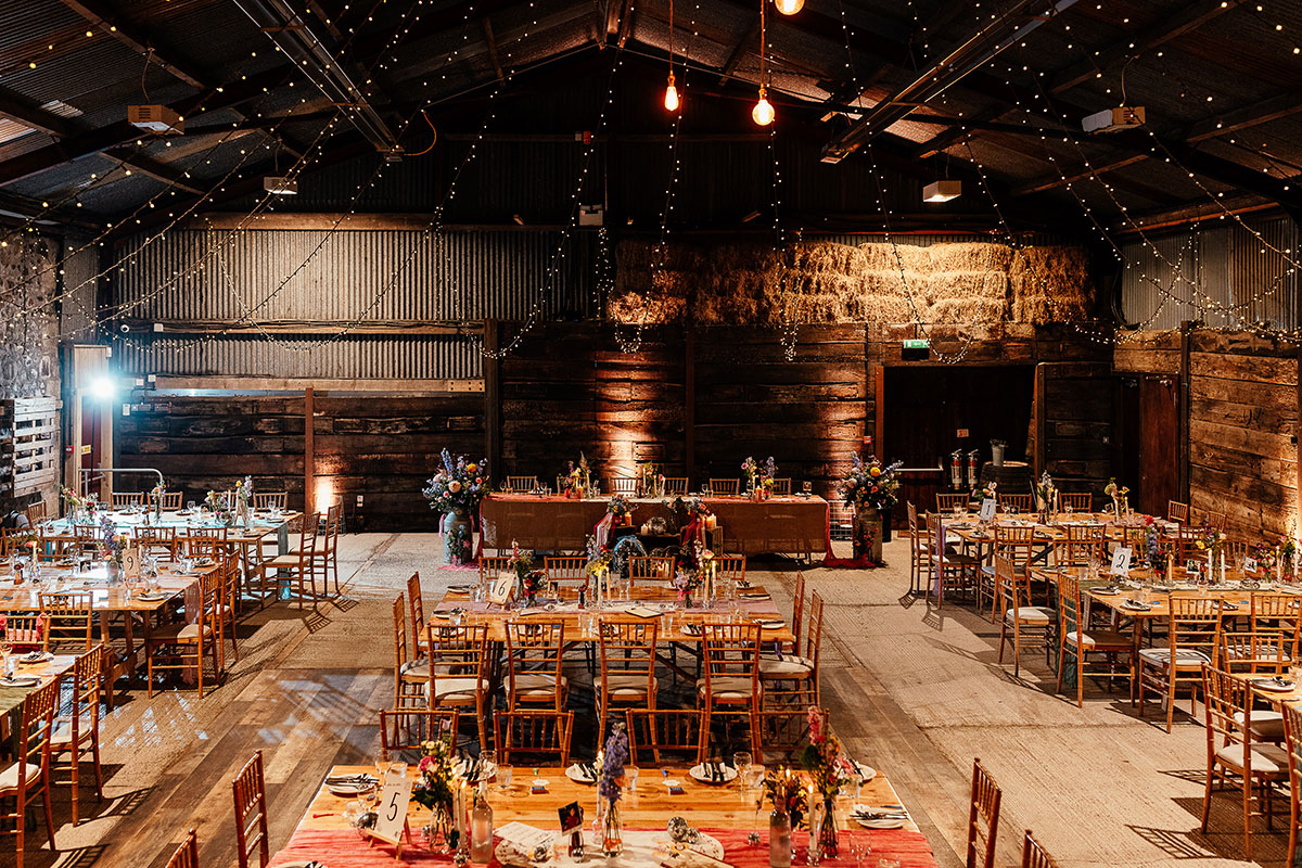 A wide view of a barn reception setup with wooden tables, chiavari chairs and overhead string lights.
